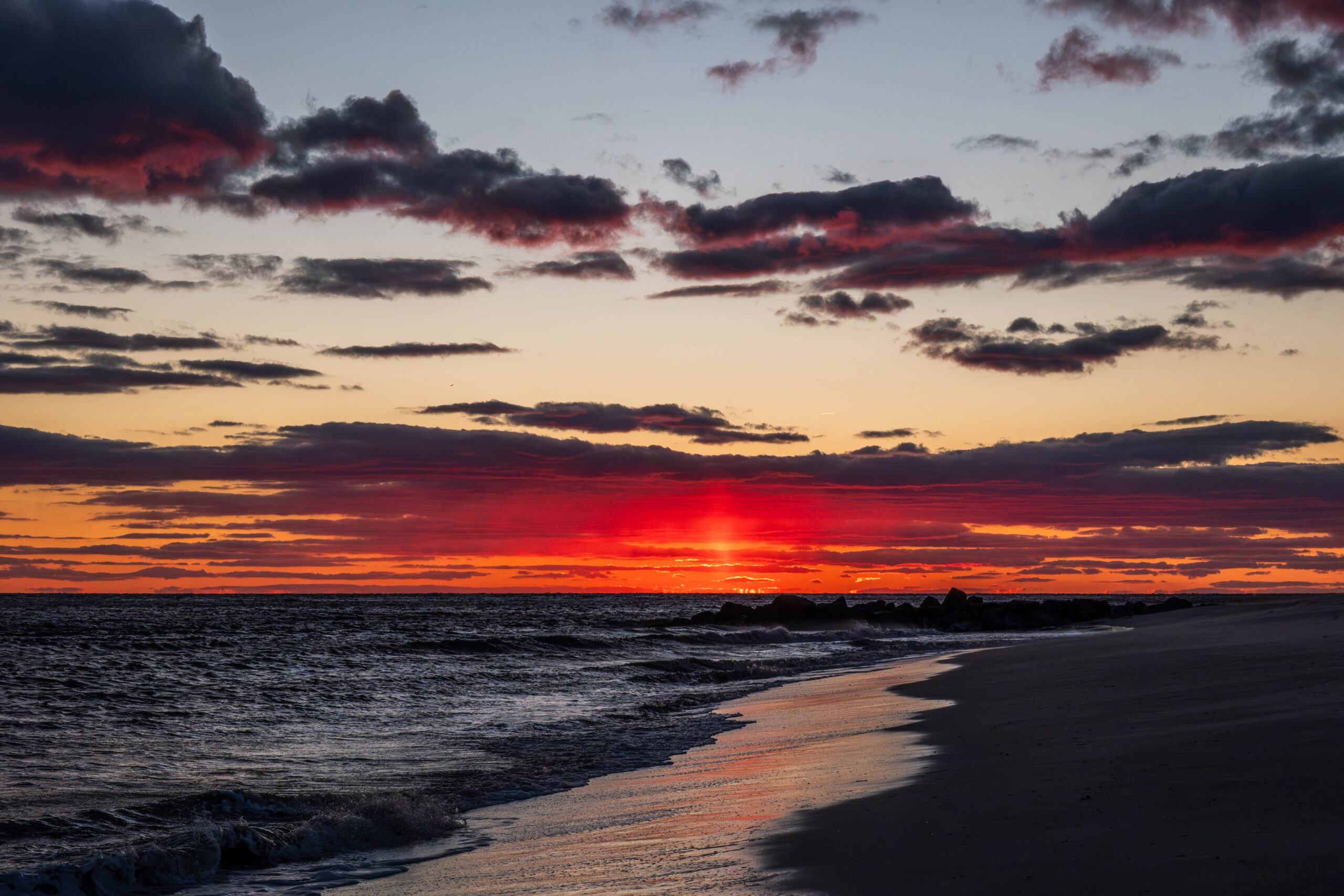 A wide view of sunset at the beach and ocean. The sun has just set, and there are dark purple and red clouds in the sky and at the horizon line. There is some clear yellow and blue sky broken up in between the clouds. Red light from the clouds is reflected in the shoreline.